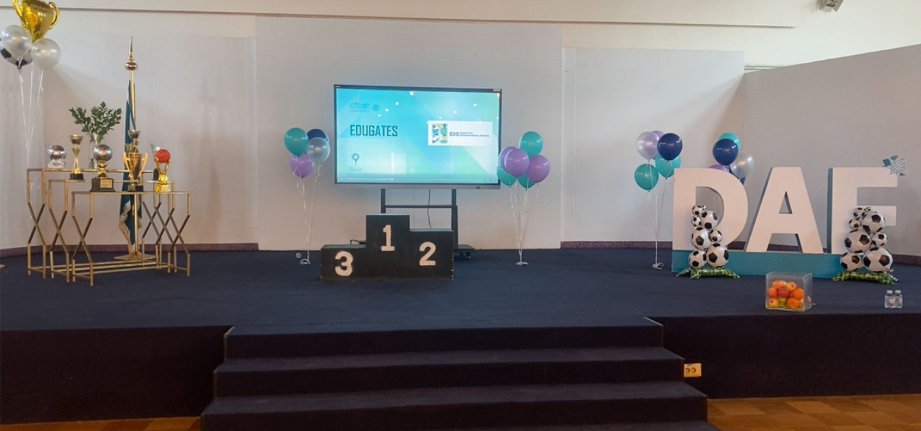 A decorated stage for the Dar Al Fikr Sports Olympics 2025 featuring trophies, a winners’ podium (1–2–3), school-colored balloons, a display screen, and large DAF letters decorated with soccer balls.